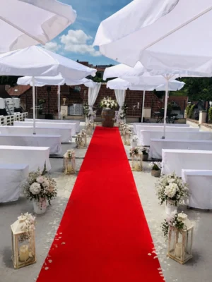 Festlich dekorierte Hochzeitslocation mit rotem Teppich, weißen Sitzbänken, Sonnenschirmen und Blumenschmuck für eine freie Trauung.