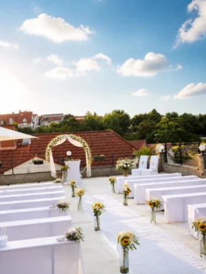 Hochzeitsterrasse mit weißen Sitzbänken, Blumenarrangements und einem festlich geschmückten Traubogen unter blauem Himmel
