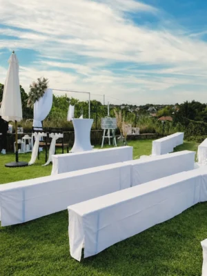 Weiße Hochzeitsbänke auf einer grünen Wiese mit dekoriertem Traubereich, Blumen und Weinbergen im Hintergrund.